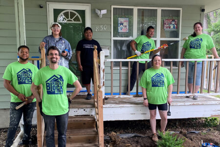 volunteers wearing Rock the Block shirts pose in front of a home with a resident of the home, one volunteer pretends to paint another volunteer