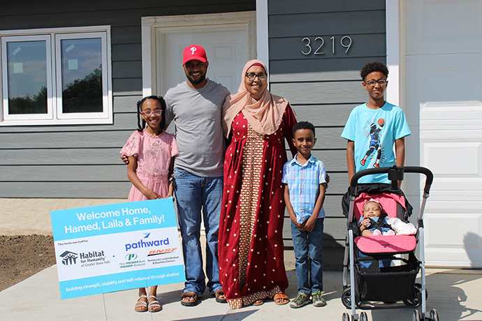 Two homeowners (Hamed and Laila) smile with their four children in front of their new home.