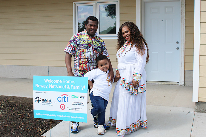 New homeowners (Neway and Netsanet) smile with their son in front of their new home.