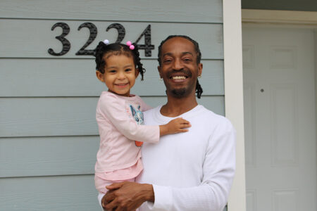 Habitat homeowner smiles with his daughter in front of their new home.