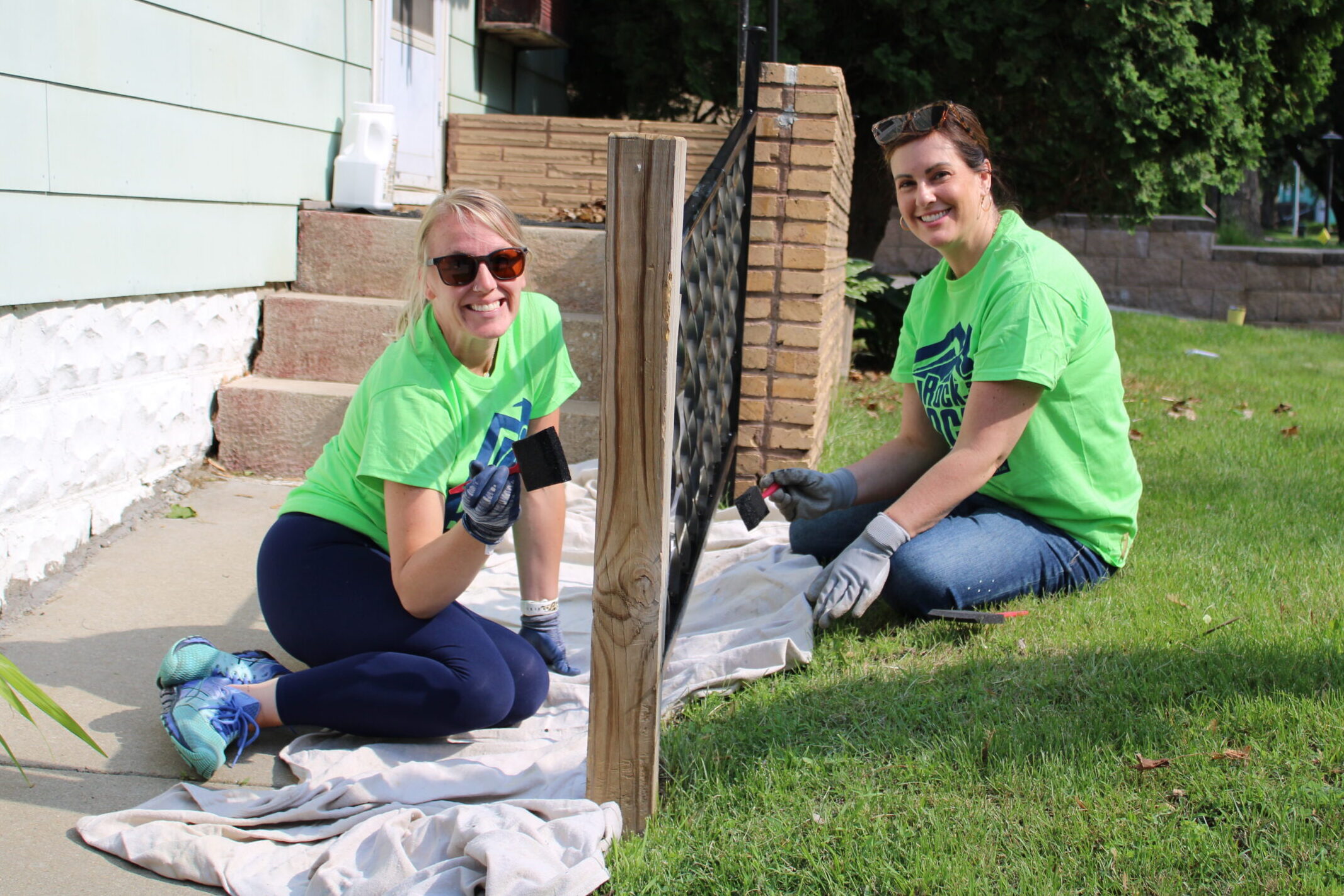 Two volunteers smile while holding paint brushes next to the porch railing they're painting.