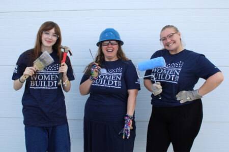 Habitat team members Emma, Brittany and Samantha pose with tools and paint brushes while wearing the 2025 Women Build t-shirt.