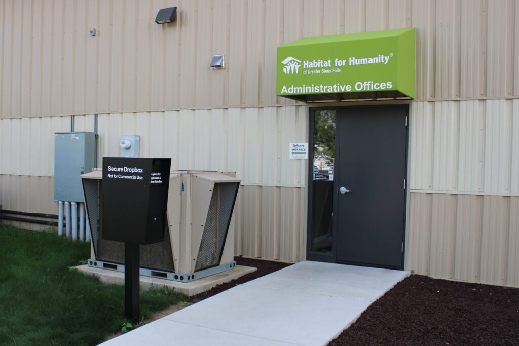 Outside of the Habitat for Humanity Greater Sioux Falls administrative offices. There is a green awning that says "Administrative Offices" with the Habitat logo. There's a tall black dropbox that says "Secure Dropbox: Not for Commercial Use".