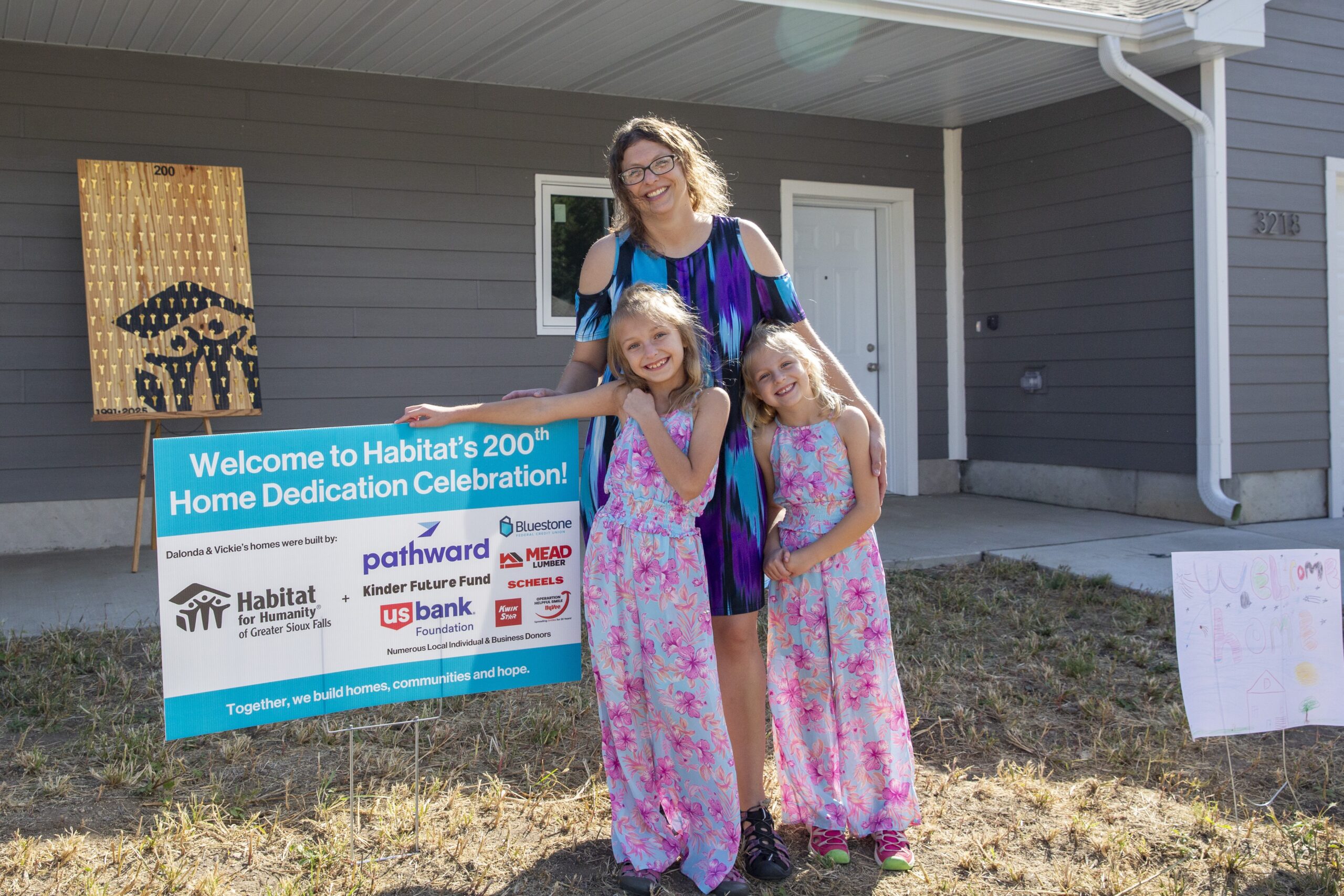 New homeowner Dalonda and her daughters stand next to the sponsor sign. It reads "Welcome to Habitat's 200th Home Dedication Celebration! Dalonda and Vickie's homes were built by: Habitat for Humanity of Greater Sioux Falls, Pathward, Kinder Future Fund, U.S. Bank Foundation, Bluestone Federal Credit Union, Mead Lumber, Scheels, Kwik Star, Hyvee Operation Helpful Smiles, Numerous Local Individual and Business Donors. Together, we build homes, communities and hope.