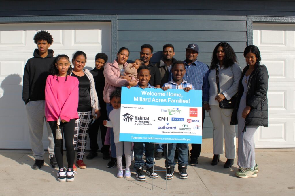 The four families stand smiling around a sign that says "Welcome Home Millard Acres Families! These homes were built by: Habitat for Humanity of Greater Sioux Falls + The Bancorp, CHN | Raven, Pathward, Wells Fargo, U.S. Bank, First PREMIER Bank & PREMIER Bankcard, Citi, and numerous local individual and business donors. Together, we build homes, communities and hope."