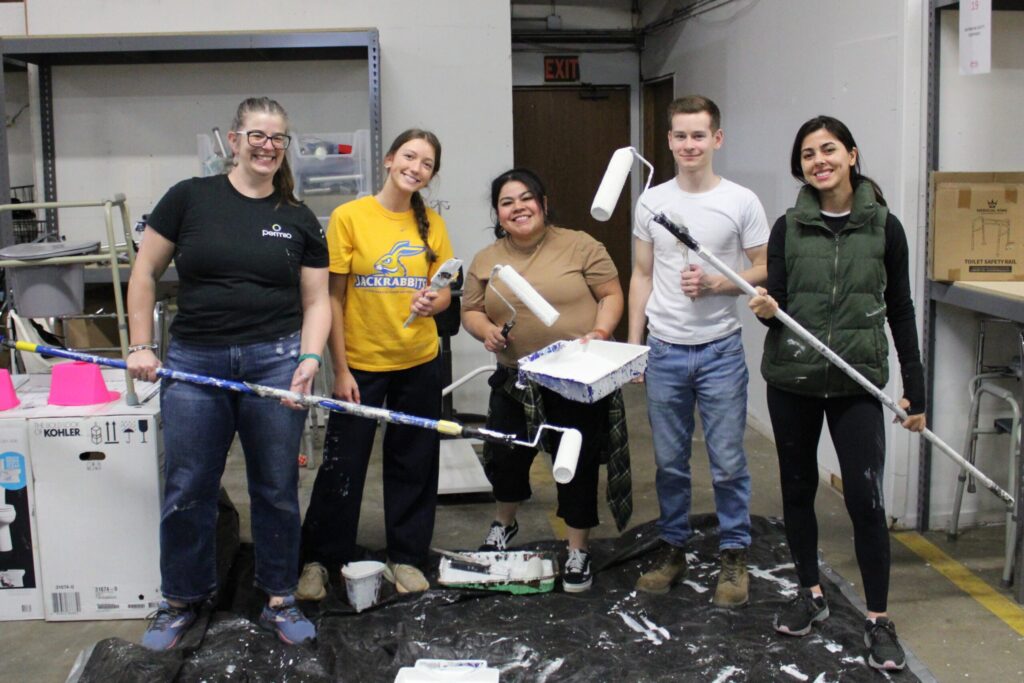 Volunteers from ISG smile with their paint supplies in the Owen's Outfitters warehouse.