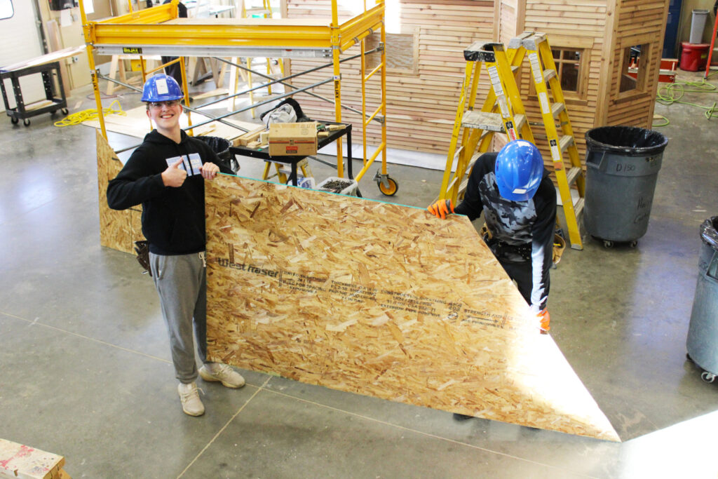 CTE Academy students carry a piece of plywood while wearing safety glasses and hardhats. One student is smiling and giving a thumbs up.