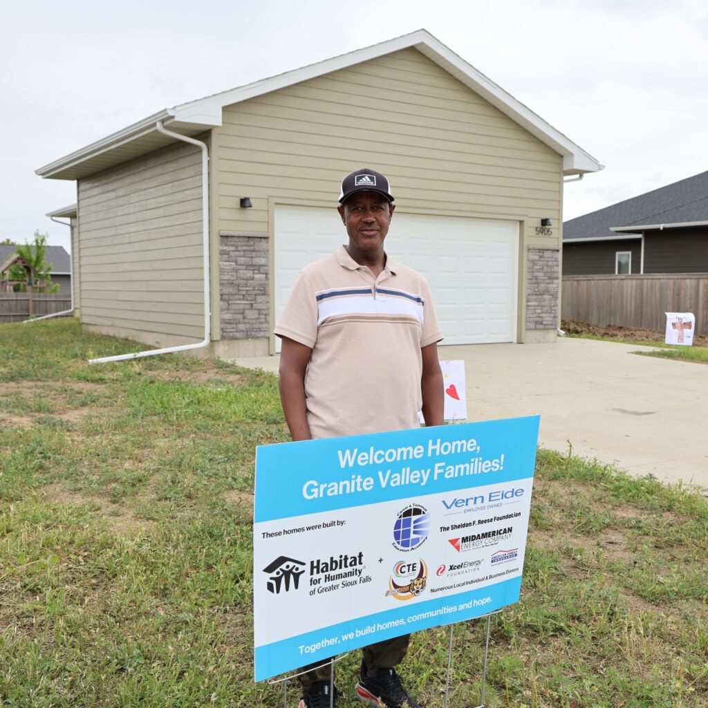 Fantahun smiles in front of the welcome home sign saying "Welcome Home, Granite Valley Families! These homes were built by: Habitat for Humanity of Greater Sioux Falls + Career & Technical Education Academy, Harrisburg High School CTE, Vern Eide, The Sheldon F. Reese Foundation, MidAmerican Energy Company, Excel Energy Foundation, American Family Insurance and numerous local individual and business donors. Together, we build homes, communities and hope."