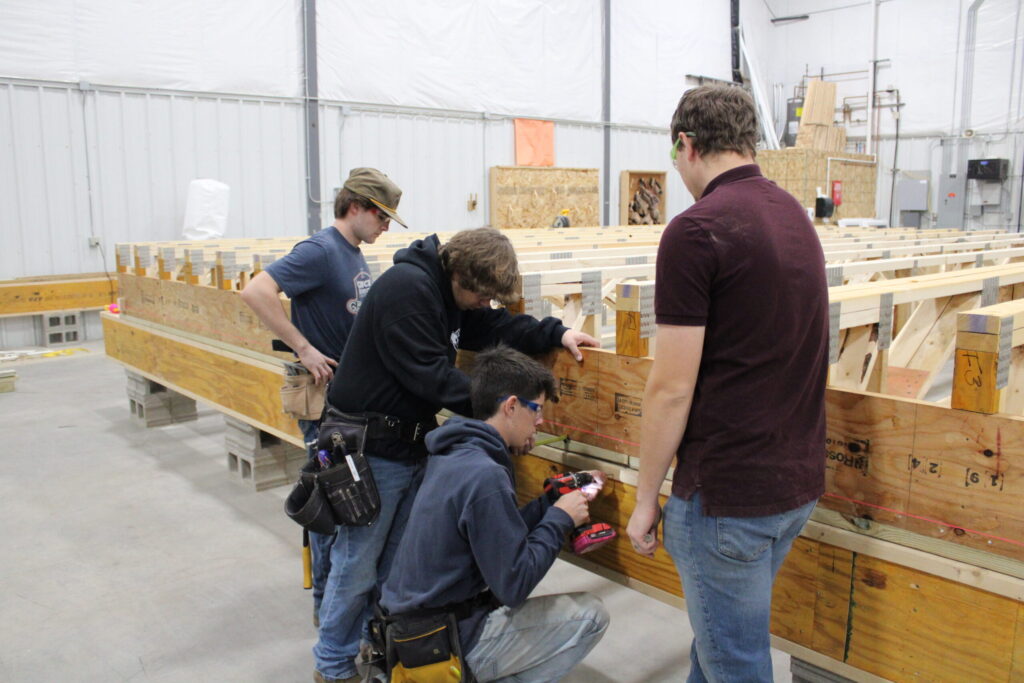Harrisburg CTE students work on the floor trusses of their Habitat home wearing safety glasses and tool belts. One student is using a measuring tape and another is using a drill.