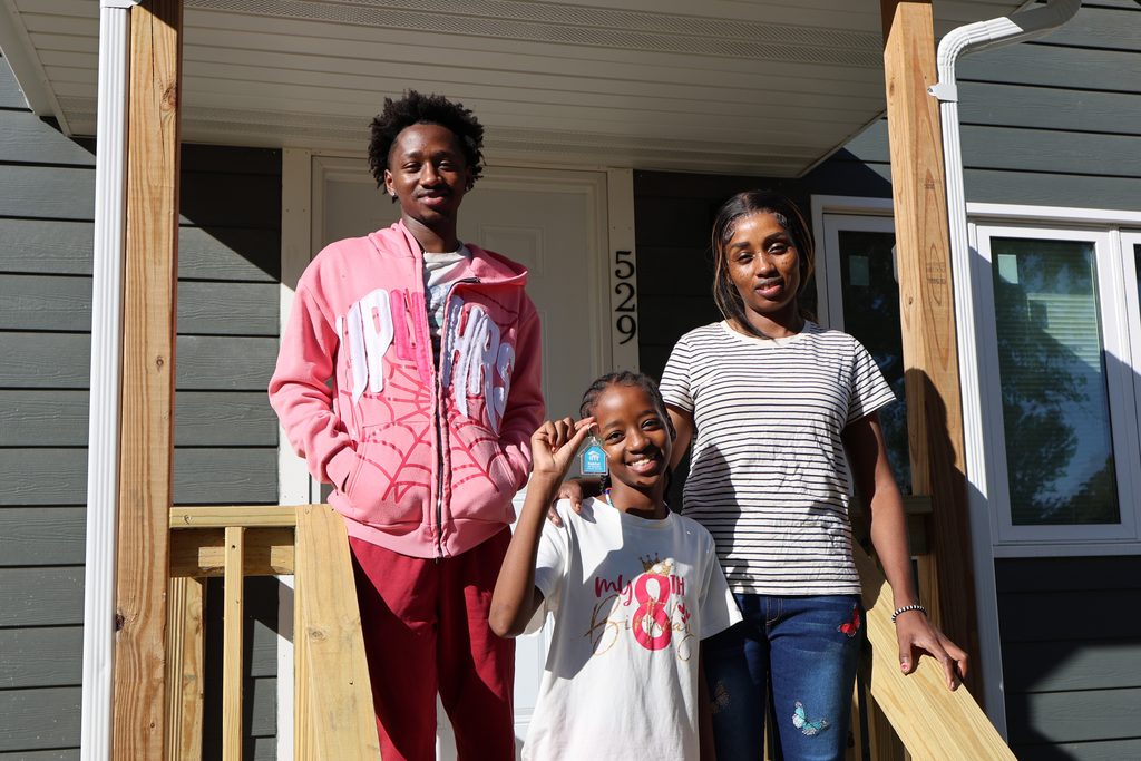Adde and her two children smile in front of their new home on their dedication day. Adde's daughter holds up a key with a Habitat house keychain.