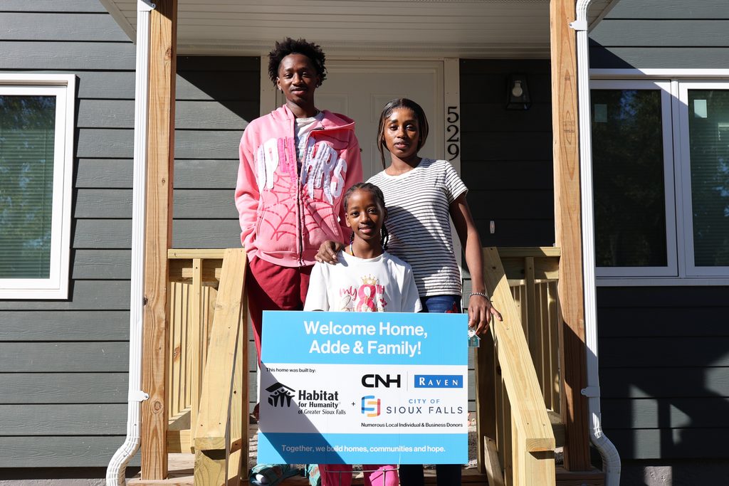 Adde and her children smile in front of their new home while holding a sign that says "Welcome Home, Adde & Family! This home was built by: Habitat for Humanity + CNH | Raven, City of Sioux Falls, Numerous Local Individual and Business Donors. Together, we build homes, communities and hope."