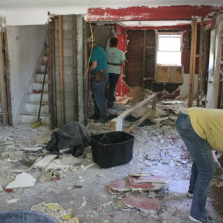 Volunteers work to take down drywall and collect it. The floor is covered with drywall pieces and insulation.