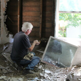 A man crouches down to work on exposed walls. The window is taken out of it's frame to ventilate the space. The floor is piled with drywall pieces and insulation.