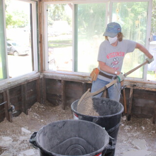 A woman wearing a safety mask shovels drywall pieces and old insulation into a trashcan.