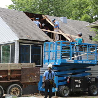 The two dormers were removed from the roofline with help from our fantastic Master's Builders. There is a hole in the roof of the house. Men are standing in the hole lifting up beams of wood to create a new roofline.