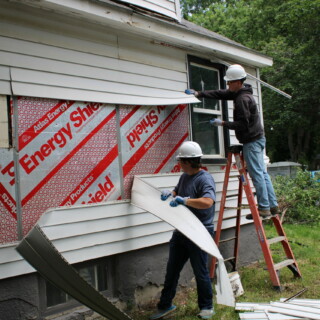 Two volunteers pull siding from the exterior of the house.