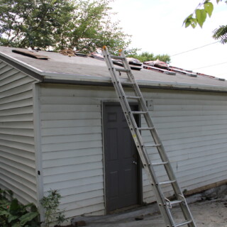 Woods Roofing, Inc. generously donated their services to shingle the house and garage for us. The garage of the house is covered with packages of shingles. Some of them have already been installed. There is a ladder leaning against the garage leading to the roof.