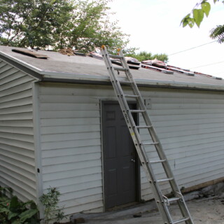 The garage of the house is covered with packages of shingles. Some of them have already been installed. There is a ladder leaning against the garage leading to the roof.