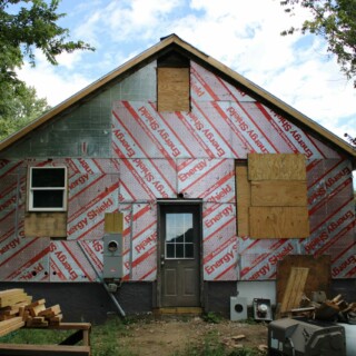 The back of the house. There is no siding. Holes from windows that were removed are covered with plywood. There is no longer a back deck, and the siding is removed leaving the energy shield exposed.