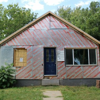 The front of the house. There is no siding leaving the energy shield revealed. A window is removed and covered with plywood. There is no longer a porch with stairs.