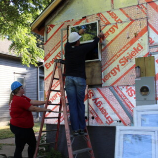 Goodbye, plywood. Hello, new windows! A man works on the window of the house. A woman is holding his ladder secure. New windows are leaning against the side of the house.