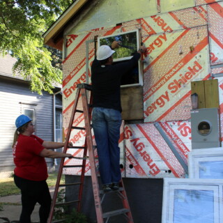 A man works on the window of the house. A woman is holding his ladder secure. New windows are leaning against the side of the house.