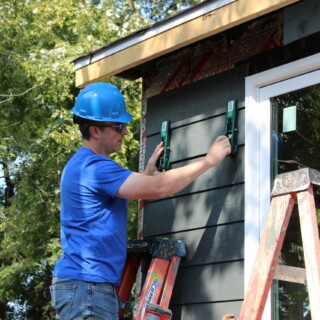 A man works to put up a piece of grey siding on the house. He's standing on a ladder.