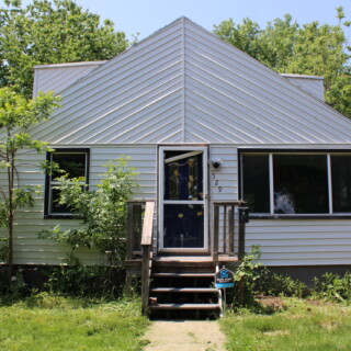 The front of the house had a broken front door, unusual roof line and overgrown landscaping. The front exterior of the rehab house before Habitat begins work. The home is white with brown trim. There is a small porch with stairs, a broken front door covered in board game money and overgrown bushes.