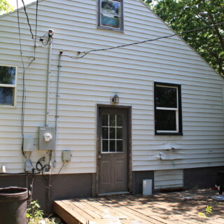 The back of the house had a deteriorating deck and broken siding. The back exterior of the rehab house before Habitat begins work. The home is white with brown trim. There is large flat deck that is deteriorating and siding is warped.