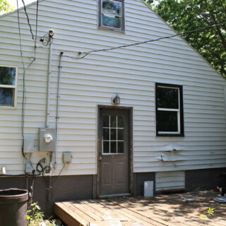 The back exterior of the rehab house before Habitat begins work. The home is white with brown trim. There is large flat deck that is deteriorating and siding is warped.