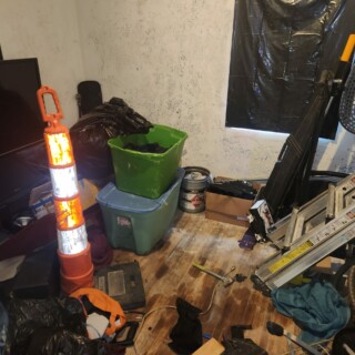 A room with grimy white walls is filled with debris. There is a window covered by a black trash bag, and the wooden floor is covered with stains.