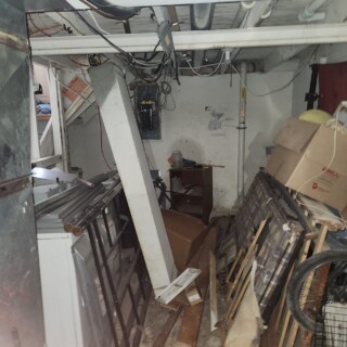 The basement was crowded with debris and had unsafe electrical wiring. A basement is filled with debris. The electrical box does not have a cover, so wires are visible.