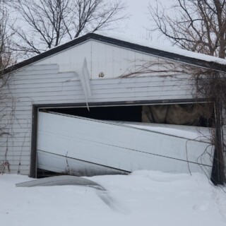 The garage door was broken and siding was peeling off the garage exterior. A garage with white siding that is peeling off of the garage. The garage door is hanging crookedly leaving large gaps where snow has collected on the inside.
