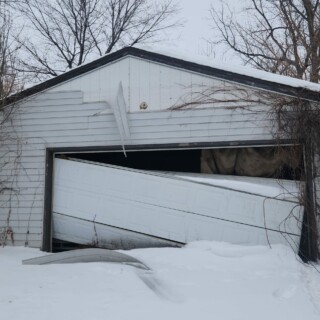 A garage with white siding that is peeling off of the garage. The garage door is hanging crookedly leaving large gaps where snow has collected on the inside.