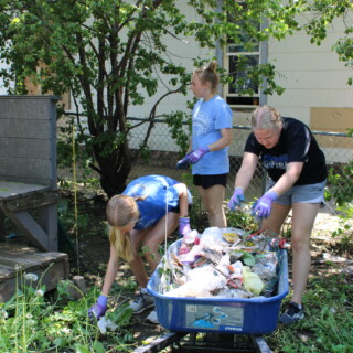 The first thing we had to do was clean up all the trash that was strewn throughout the yard. Thank goodness for rubber gloves! Three young women pick up trash in the yard and add it the pile already collected in a wheelbarrow.
