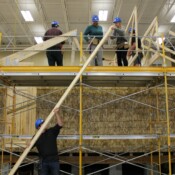 A student hands up a board to other students that will be used to truss a home. The home is partially constructed with scaffolding constructed around it. Students are wearing hard hats and tool belts.