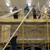 Construction students at CTE Academy work together to add trusses to their Habitat home. A student hands up a board to other students that will be used to truss a home. The home is partially constructed with scaffolding constructed around it. Students are wearing hard hats and tool belts.