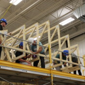 Construction students at CTE Academy work together to add trusses to their Habitat home. A CTE Academy instructor teaches students how to properly secure the trusses. They are all wearing hard hats and are standing on top of the scaffolding.