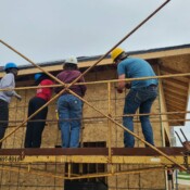 People standing on scaffolding secure a piece of wood to the garage. They're all wearing hardhats.