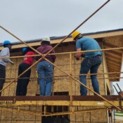 Rebecca, the future homebuyer, and her daughter work with Habitat staff and volunteers to build the garage. People standing on scaffolding secure a piece of wood to the garage. They're all wearing hardhats.