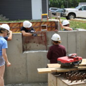 A group of volunteers with hardhats and hammers work on the foundation of the home. They are being instructed by Jolynn, Habitat's construction site supervisor.