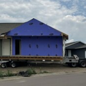A large truck carrying a home backs the home up in line with the foundation. The home is sided except where the garage will be added.