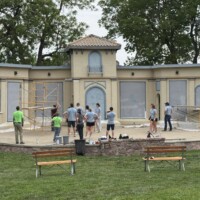 The bandshell is yellow and gray with patch spots. Volunteers stand listening to instructions for painting.