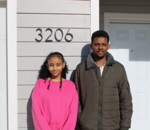 Tesfaye and one of his children smile in front of their new Millard Acres twin home on their home dedication day.