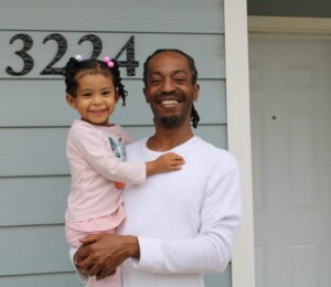 Josiah smiles while holding his daughter outside of their new home.